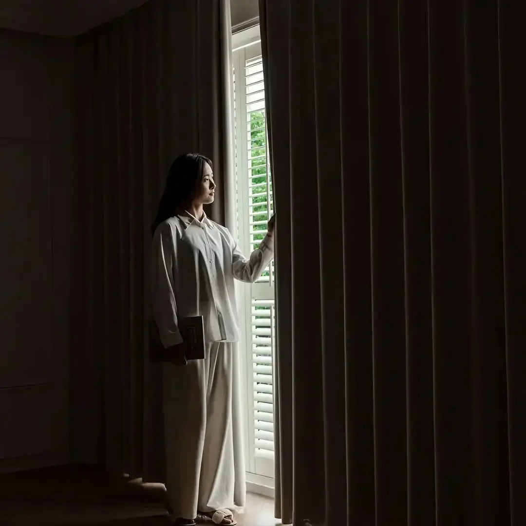 Pessoa abrindo uma fresta entre os painéis fechados da Cortina Valencie bege em quarto escuro