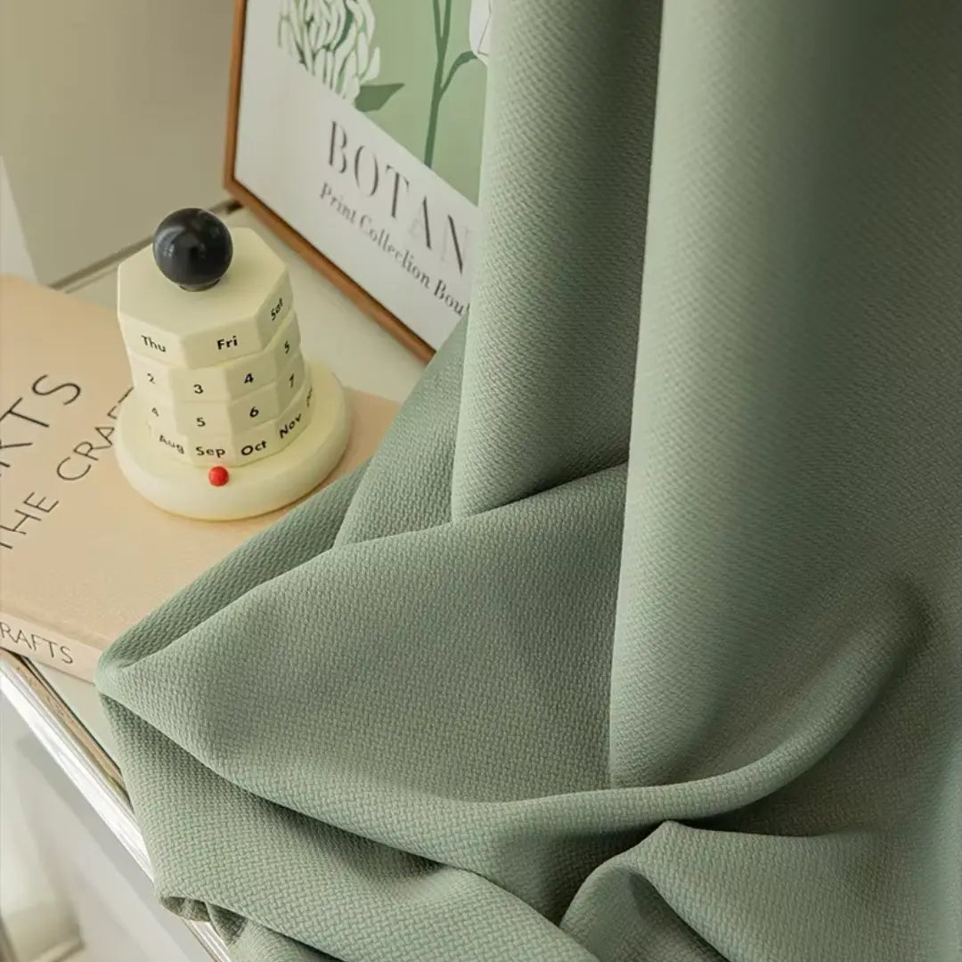 Tecido verde da Cortina Valeriana drapeado sobre uma superfície, ao lado de livro e caneca