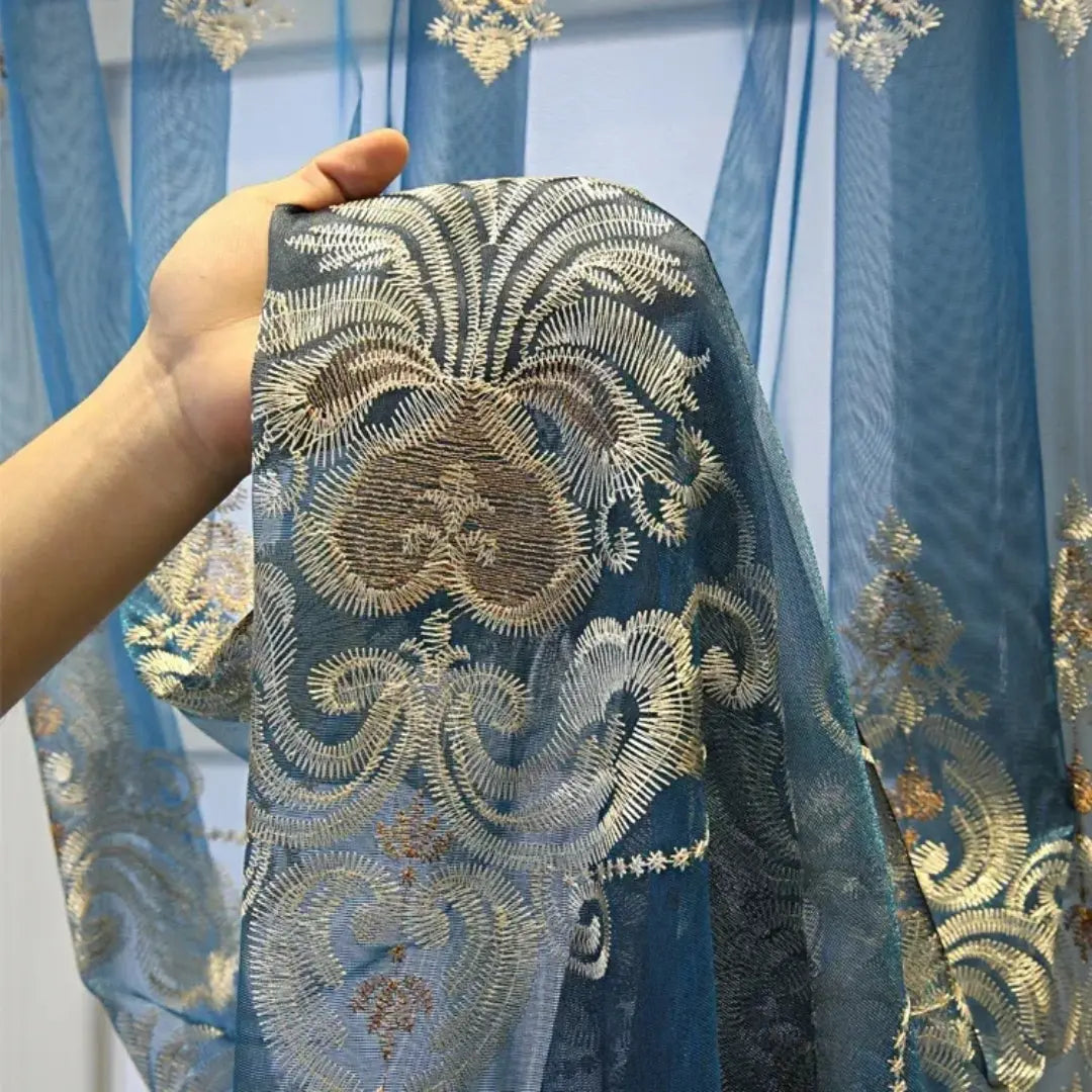 Mão exibindo o bordado floral azul e prateado da cortina Éclat D'Or sobre o voil azul