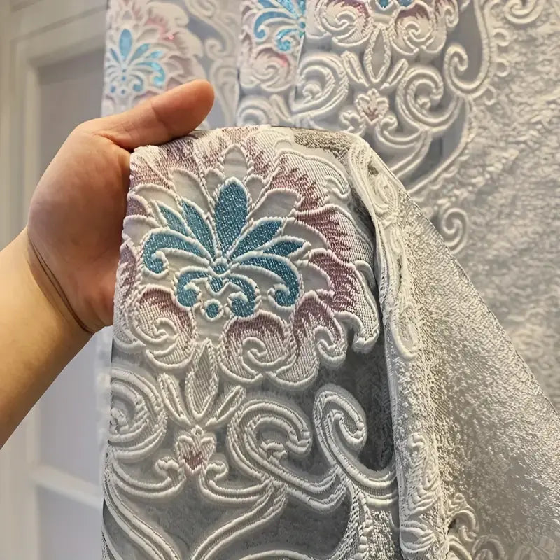 Mão segurando o tecido da Cortina Winterfell com flor turquesa e rosé entre arabescos bordados