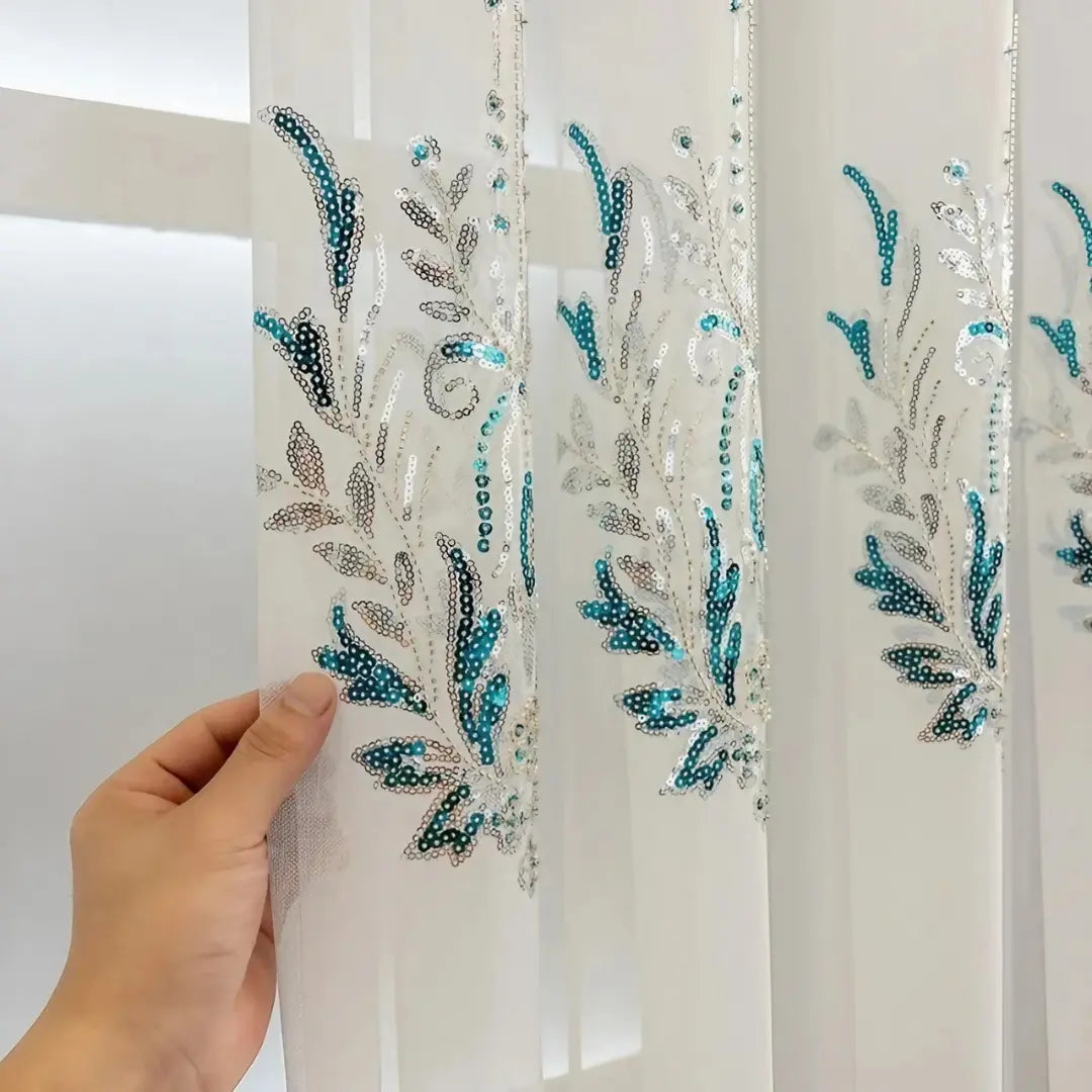 Mão exibindo o bordado azul-turquesa e prateado da Éléonore sobre o voil branco translúcido