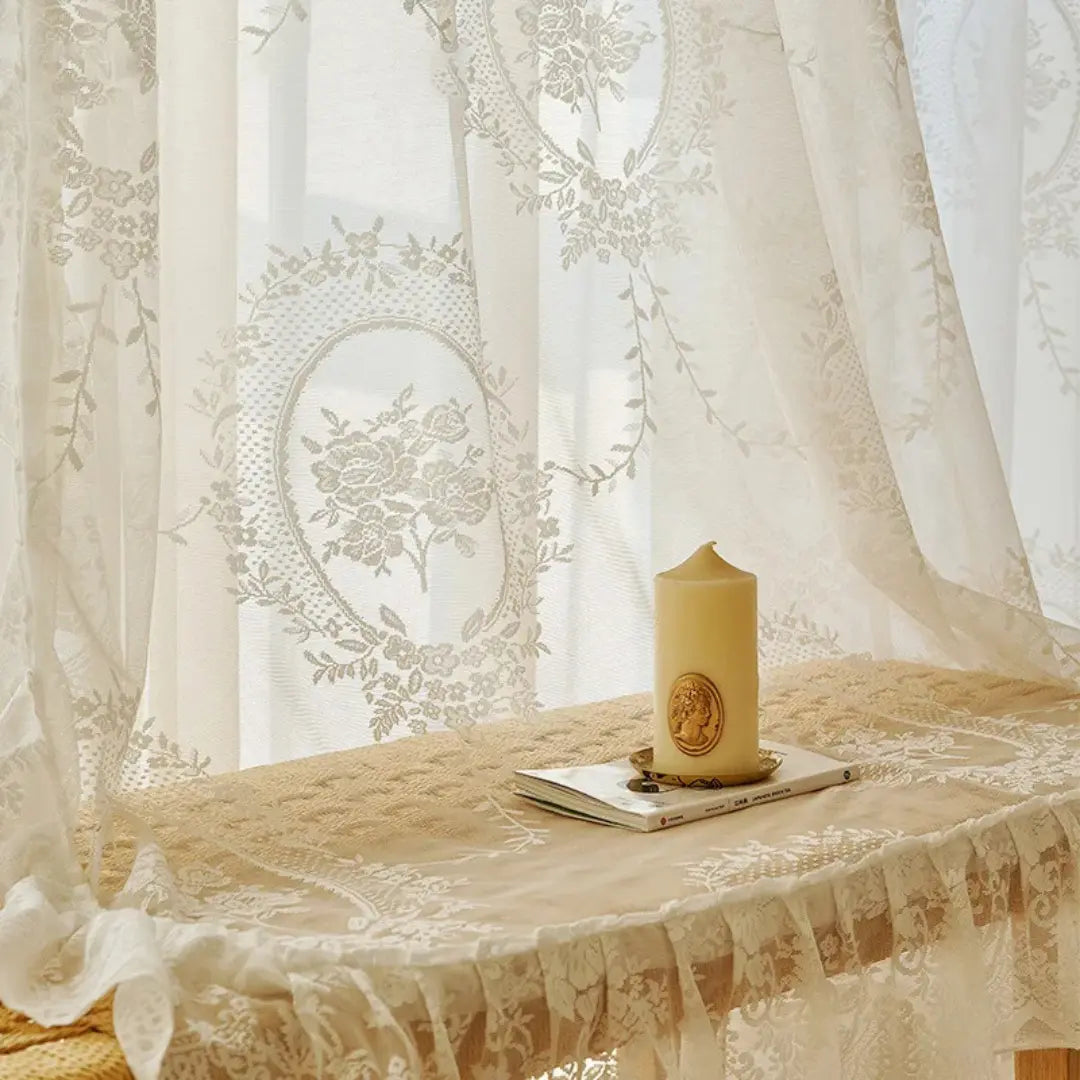 Renda bordada da Cortina Valerys drapeada sobre mesa, com vela e livro em composição clara